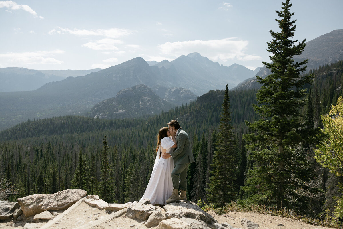 Bride and groom kissing on a cliffside in Rocky Mountain National Park with mountain peaks in the background, captured by a destination elopement photographer.