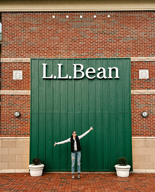 A woman stands outside an L.L. Bean store, smiling and looking at the entrance.