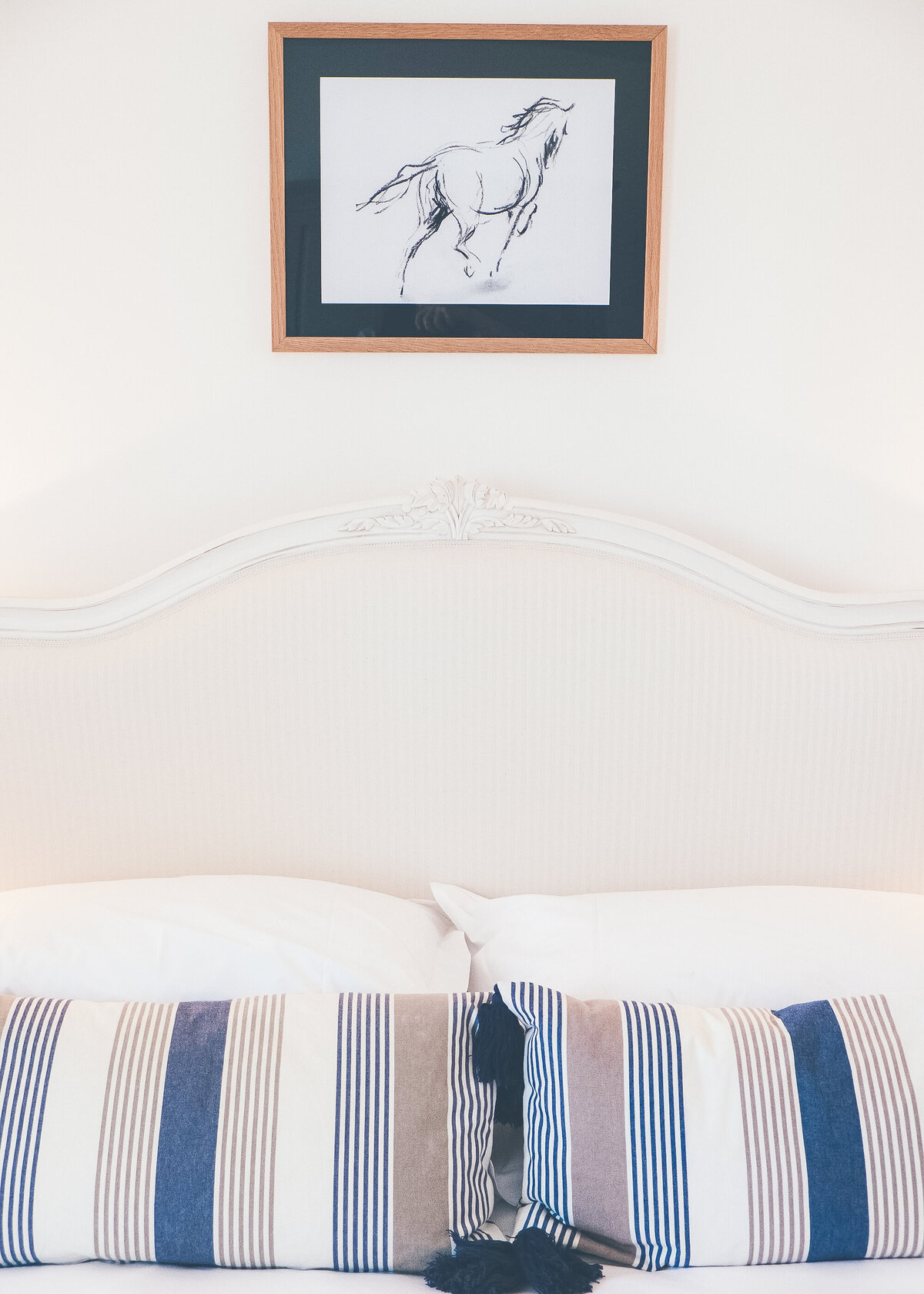 A white bed is styled by an interior designer with white and blue striped cushions under a horse line portrait.