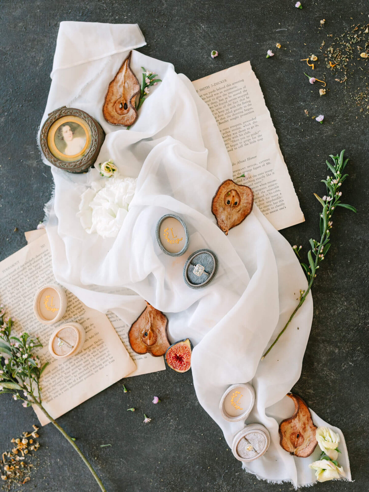 Artistic flat lay of vintage-style rings in ornate boxes surrounded by dried fruit slices, flowers, and book pages.