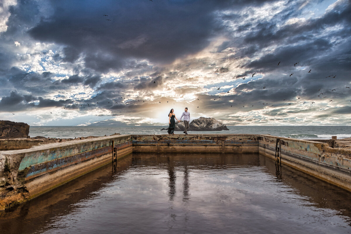 Philippe Studio Pro, a Sacramento wedding photography studio, captures a couple walking across Sutro Bath in the San Francisco Bay area with dramatic sky, clouds and sunset in the back while holding hands.  Photo creatively captured and edited by philippe studio pro, from Sacramento.