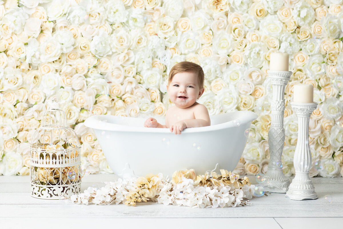 Dallas milestone photographer captures a joyful baby in a vintage-style white tub surrounded by soft cream florals, decorative candles, and bubbles for a charming post-cake smash bath session.