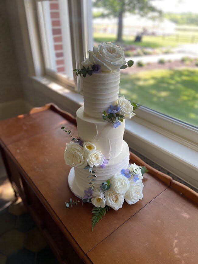 A white wedding cake sits on a wooden table in front of a window. The cake has three tiers with alternating textured buttercream. The top and bottom have line texture, and the middle has a a subtle spackle layering texture. There's clusters of fresh flowers in white and pale blue.