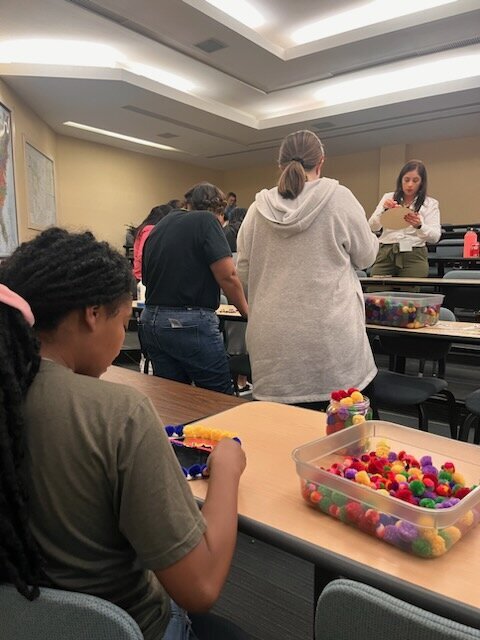 The creative journey as two women work on a wooden table, one carefully gluing vibrant pompoms around a round mirror while the other observes.