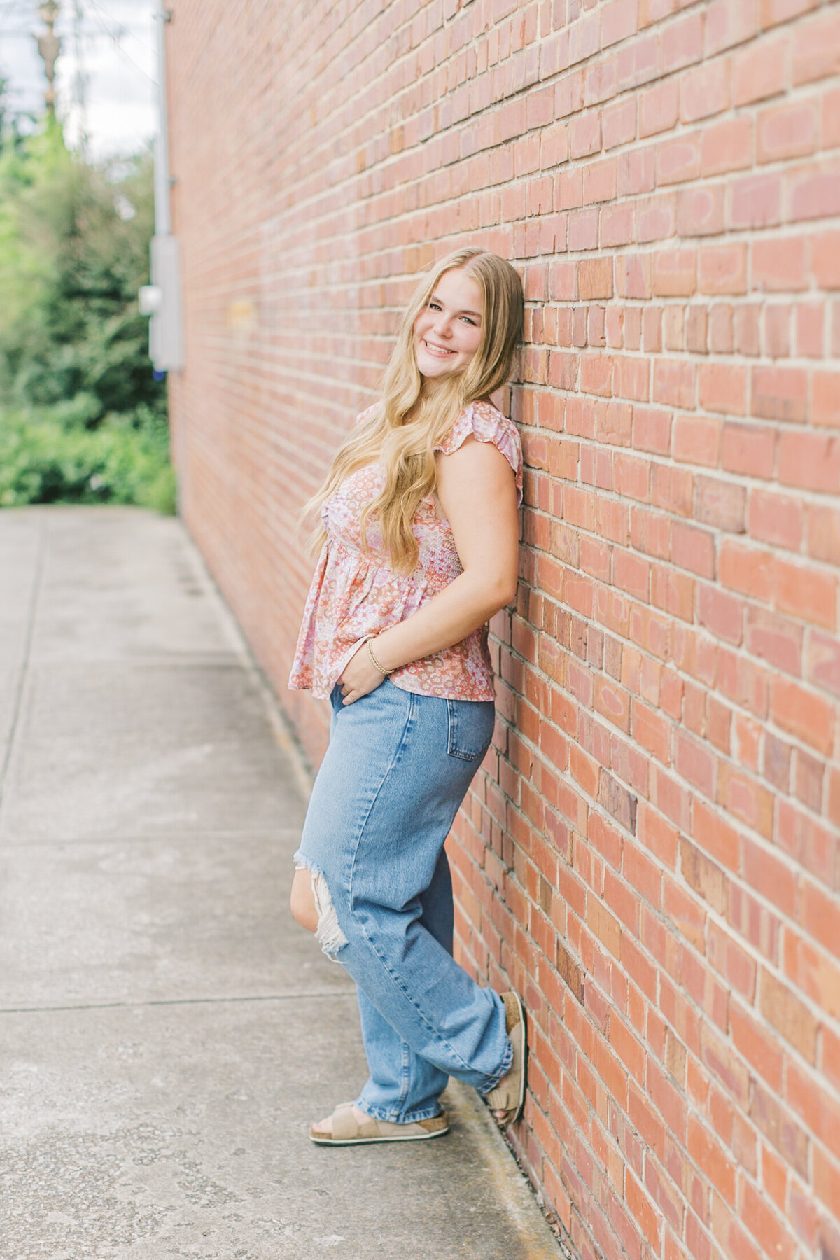 A senior girl leaning against a red brick wall, smiling confidently during her downtown session — Raleigh portrait photography.