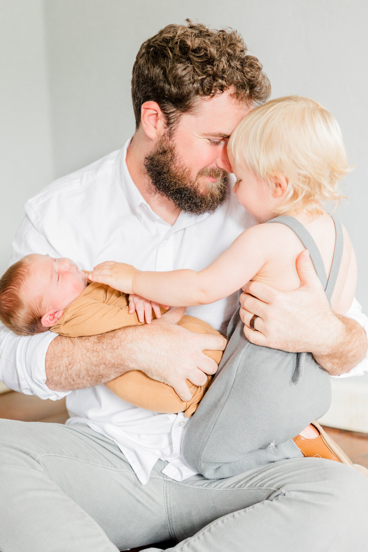 Dad sitting holding baby and toddler in his lap, cuddling toddler while toddler touches newborn