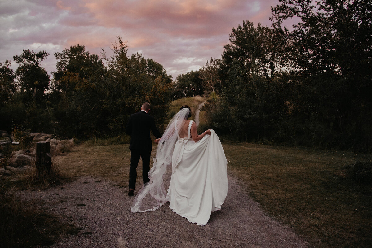 Classic and stunning bride and groom, walking along path with gorgeous pink and blue sunset in background, blue hour wedding, captured by Kelsey Vera Photography, intimate and romantic wedding photographer in Airdrie, Alberta. Featured on the Bronte Bride Vendor Guide.