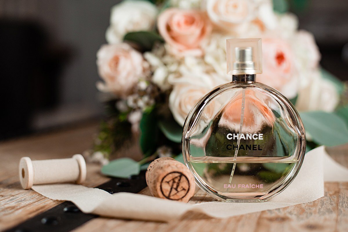 Round bottle of Chanel Chance perfume sitting on a wood table with a spool of silk ribbon and an Arrington Vineyard wine cork with a bouquet of pink roses, white roses, white hydrangea and eucalyptus in the background.