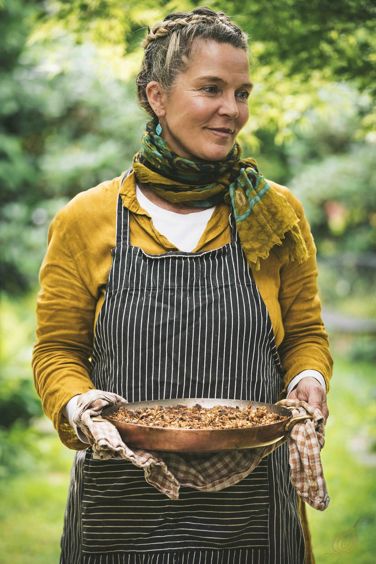 A woman in an apron stands holding a pan filled with delicious food, ready to serve a meal.