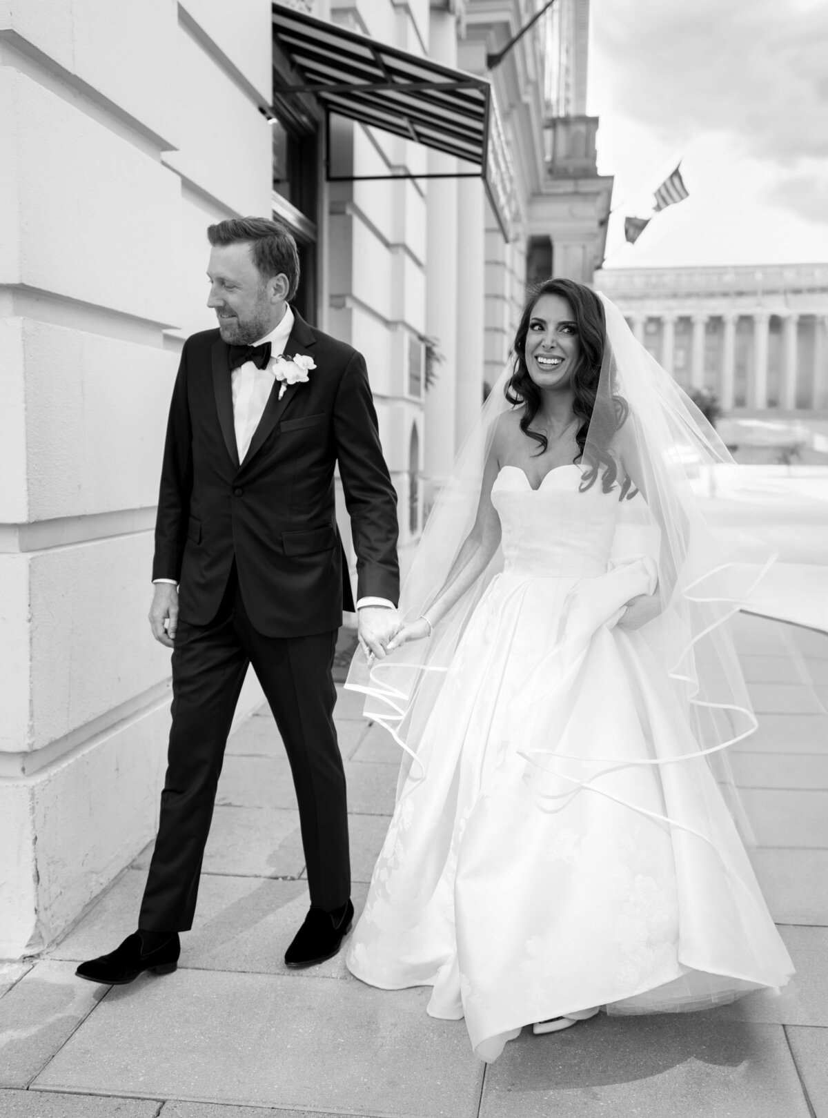 A bride and groom joyfully hold hands while walking down a city street. The bride wears a flowing white gown and veil, and the groom is in a dark suit with a bow tie and boutonniere. The background features a building and an American flag.