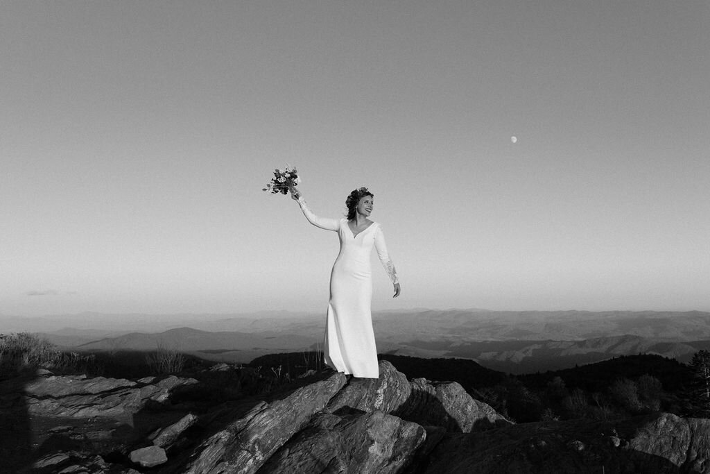 Bride with flower crown standing on a rock holding her bouquet in the air with a mountain scape behind her