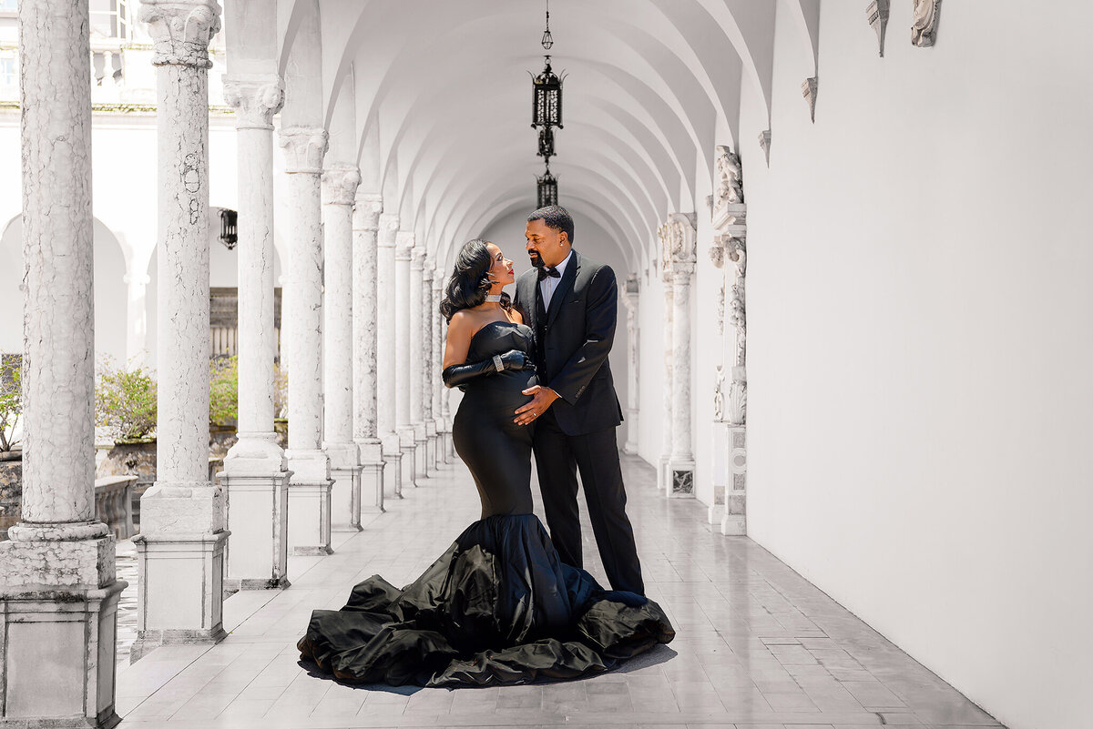 Expecting couple in formal black attire, gorgeous black fitted gown with mermaid bottom, looking at each other lovingly, standing in corrdor of white ourdoor courtyard