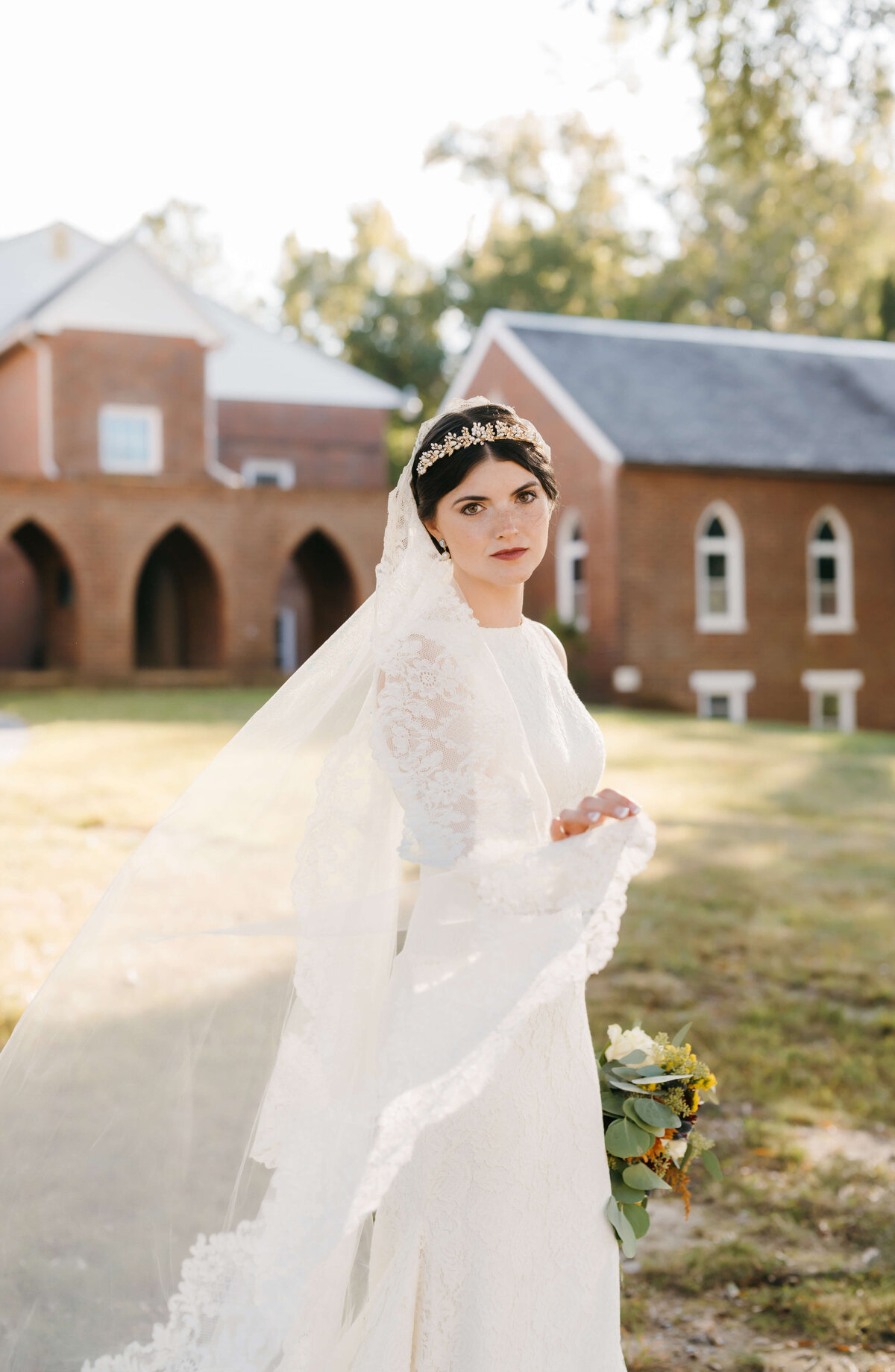 bridal portraits by Virginia wedding photographer with bride in an ethereal wedding gown and veil poses in front of a colonial styled chapel for her richmond wedding as she holds her bouquet to the side and her other hand holds her veil gently