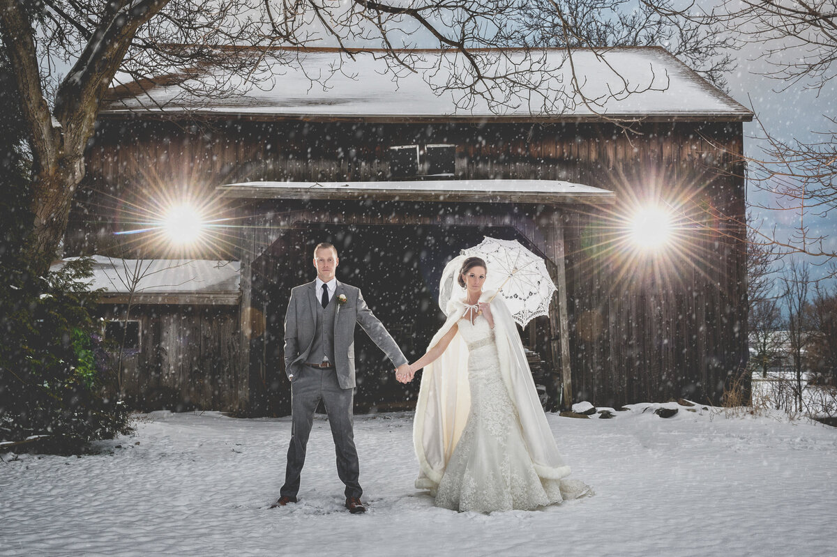 Bride and groom holding hands in the snow by barn.