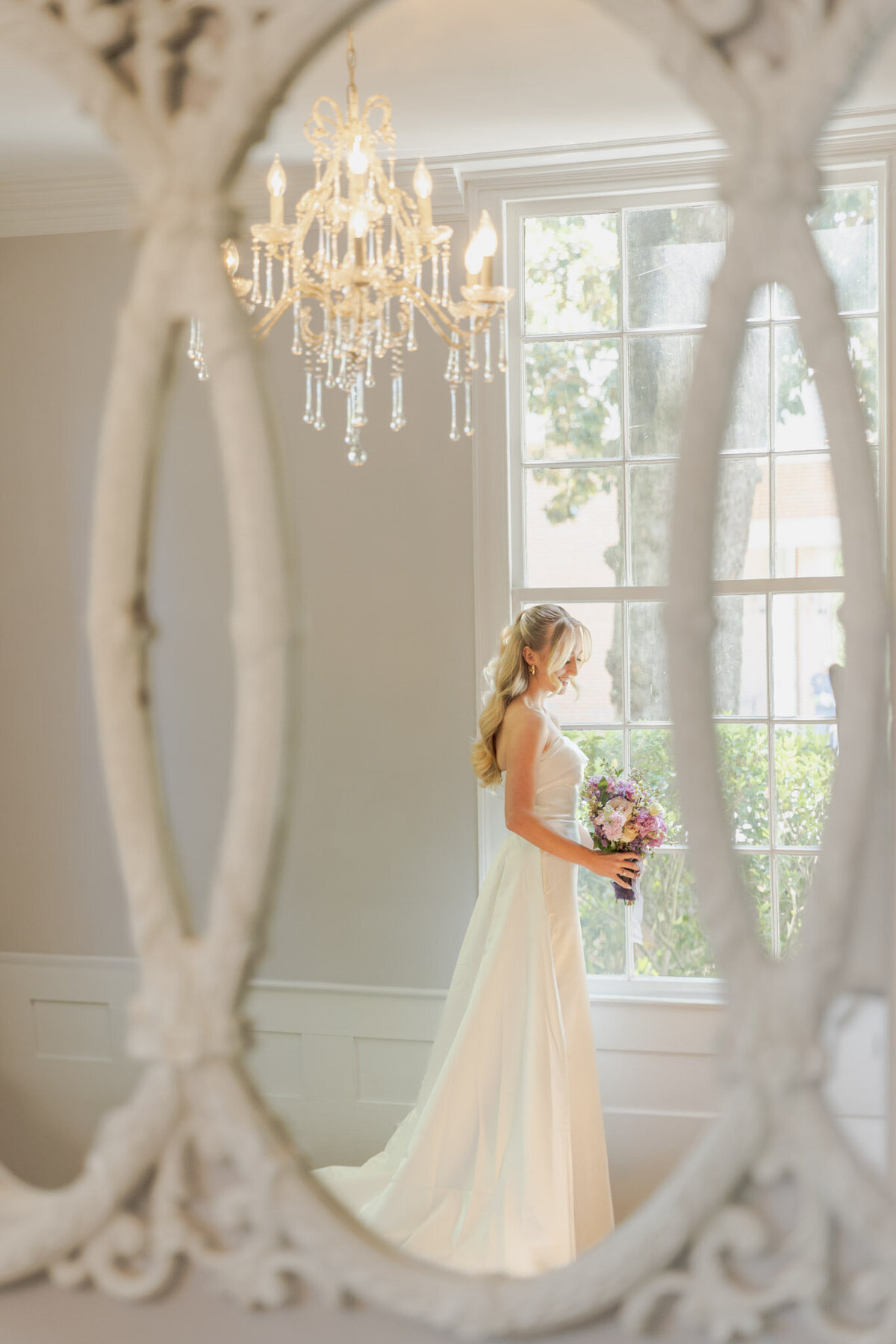 Looking out a large window, a bride is all dressed up and holding her wedding bouquet.
