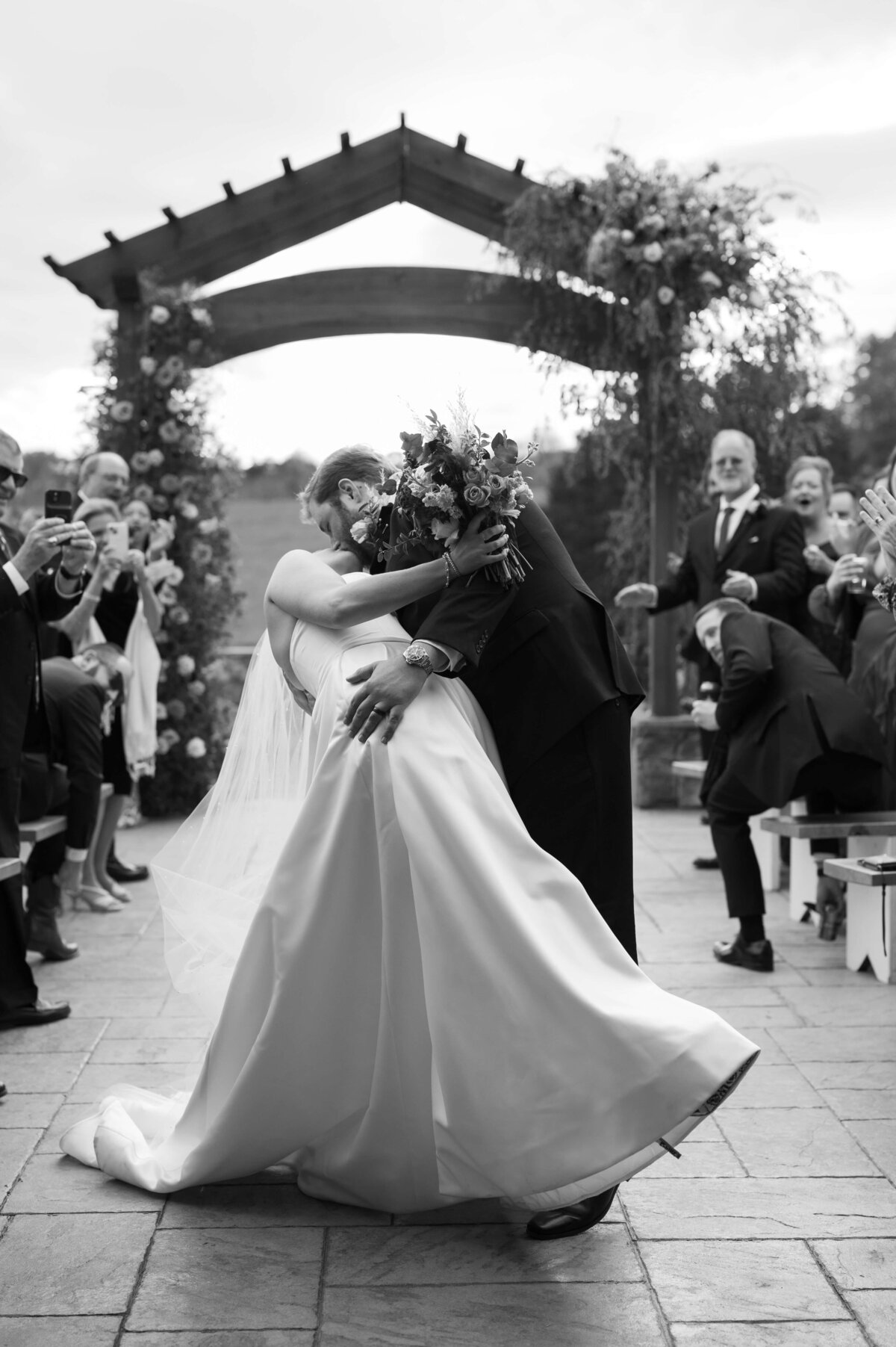black and white wedding picture with groom dipping his bride backwards at the end of the aisle as they exit their wedding ceremony with their guests standing and clapping in celebration