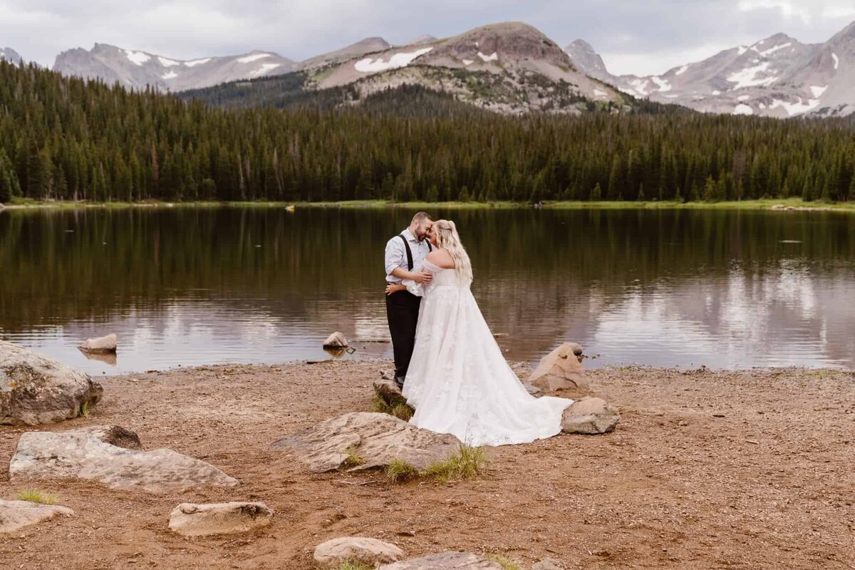 Bride and groom embrace beside Brainard Lake with dramatic mountain peaks in the background during their Colorado elopement.