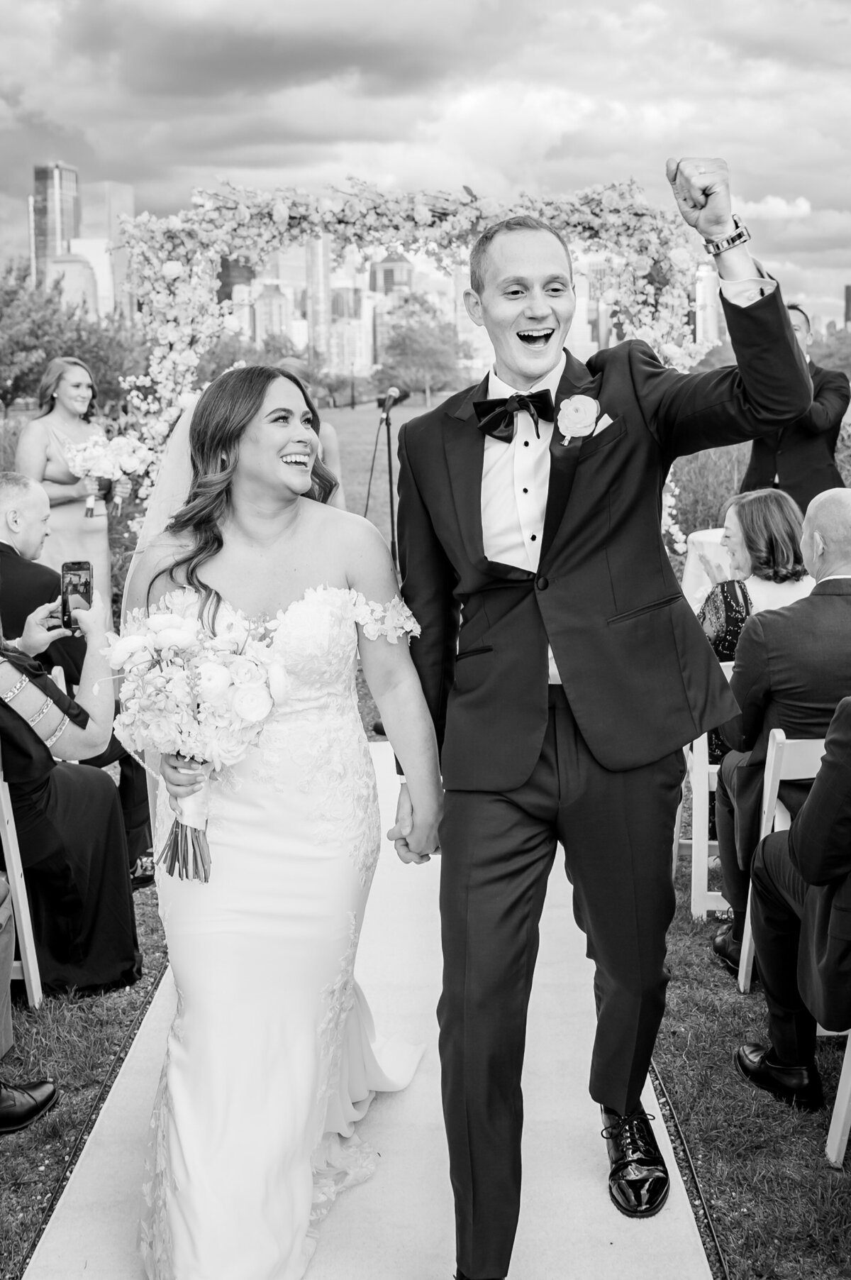 A newlywed couple walks down the aisle outdoors after their wedding ceremony at Liberty House in Jersey City.  They are holding hands and smiling. The groom raises his fist in celebration, while guests seated on either side cheer. Captured with stunning candid wedding photography, the NYC city skyline provides a breathtaking backdrop for this joyous moment.