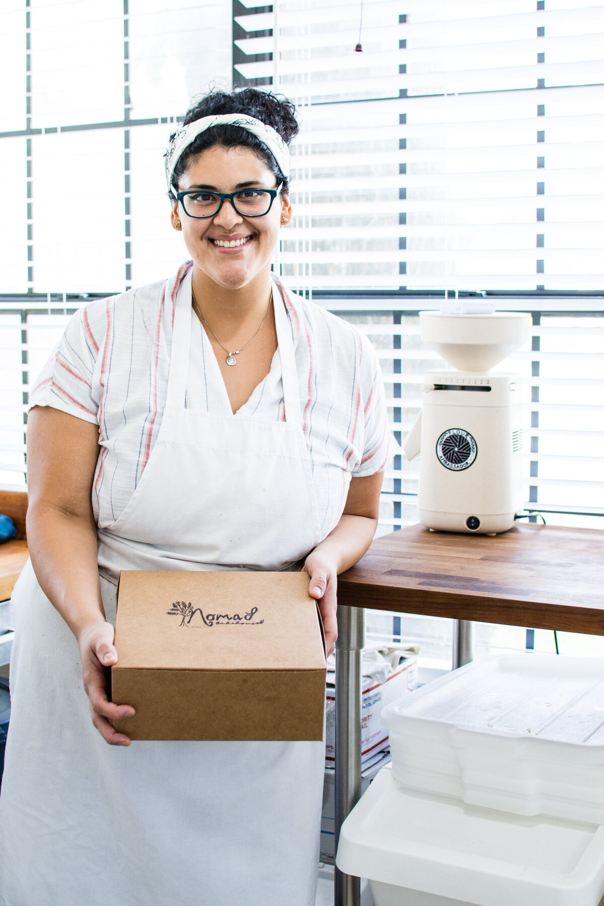 Smiling woman with dark hair and white apron holding a brown box with the words Nomad Bakehouse stamped on top