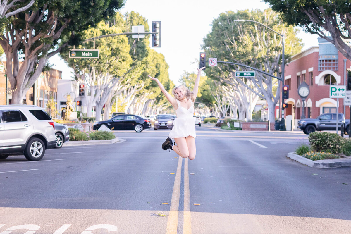 A person jumps joyfully in the middle of an empty street lined with trees. Cars are parked on both sides, and traffic signs are visible. The background shows buildings and a partly cloudy sky.