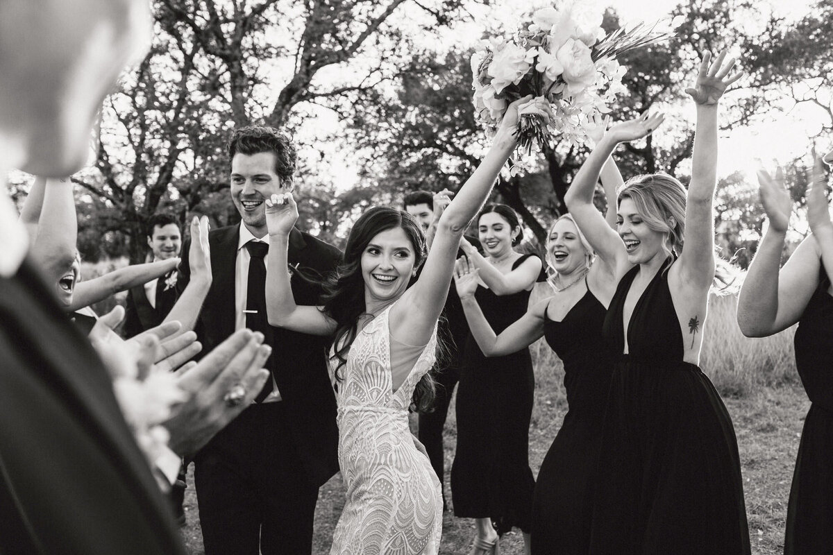 A black and white photograph of Jade and Brian on their wedding day at Mae’s Ridge in Johnson City near Austin, TX. Jade raises her arms in celebration as she and Brian walk between their friends in their wedding party who are cheering, smiling, and waving their arms. Wedding photography by Stacie McChesney of Vitae Weddings.