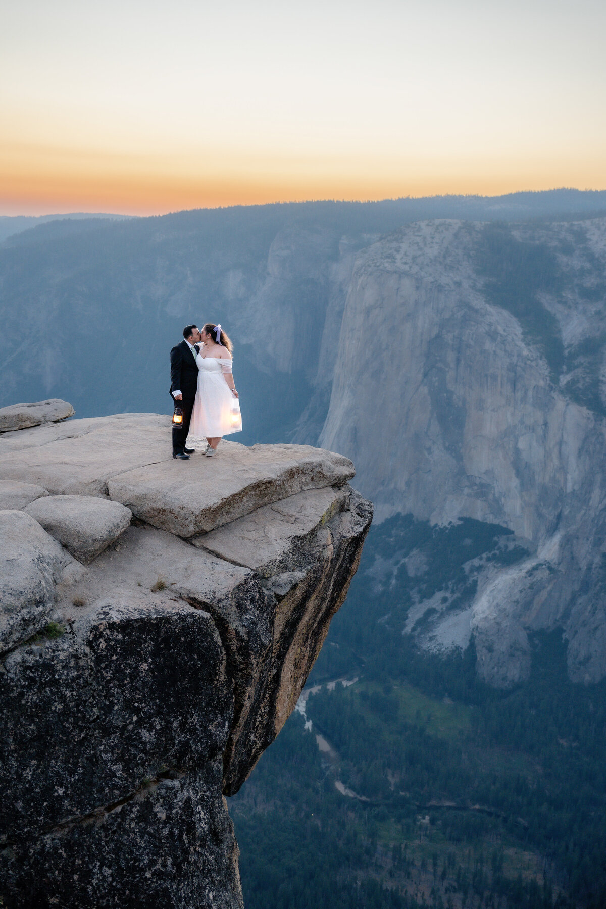 Bride and Groom eloping at Yosemite National Park Taft Point cliff during sunset holding lanterns and kissing.