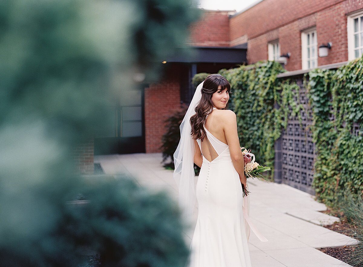 The bride, wearing a silk sheath dress and long veil looks over her shoulder at the camera in the ivy covered courtyard at Clementine Hall.
