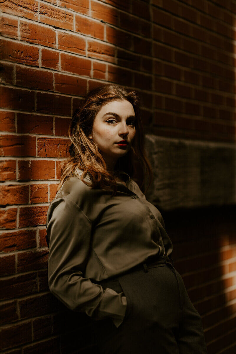 femme rousse debout, regard perçant, en tenue kaki, adossée à un mur de briques rouges, prise lors d'un shooting photo portrait à Montréal.