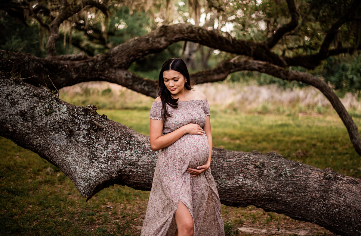 An expectant Houston area mom holds the top and bottom of her bump and looks at her shoulder while standing near an oak tree.