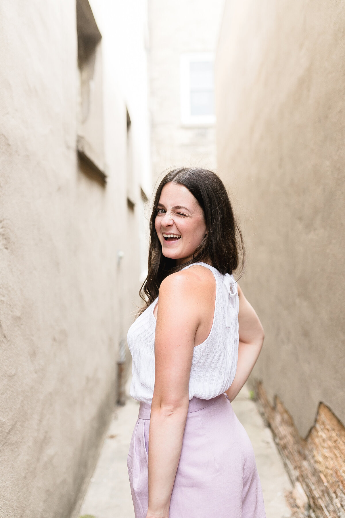 High school senior girl posing outdoors in a cream rustic alley, wearing white tank top with lavender skirt, winking at camera