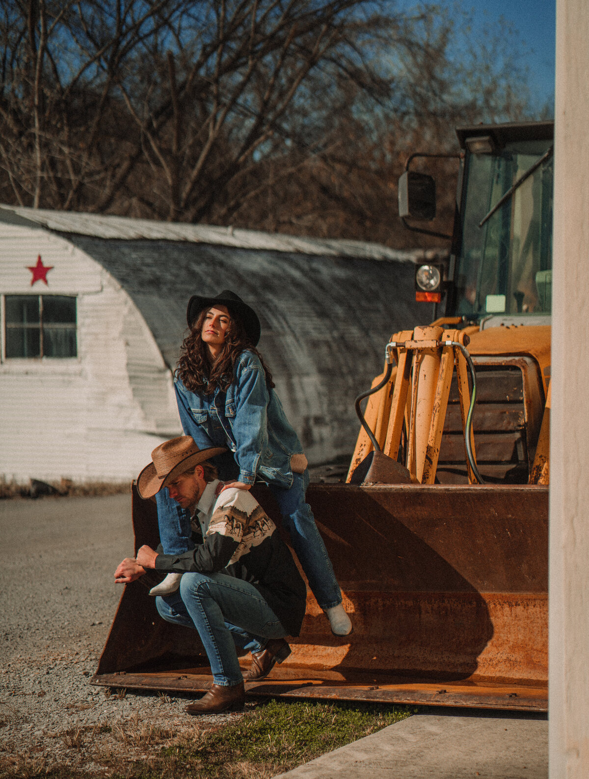 A girl in a cowboy hat sits on the bucket of a dozer while a man in a cowboy hat kneels in front of her.