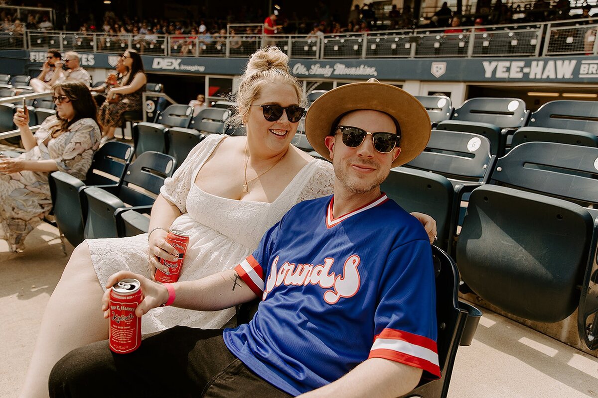 The bride, wearing a square neck lace mini dress holds a red can as she puts her arm around the groom. The groom, wearing black pants and a blue Nashville Sounds jersey and fedora smiles for the camera holding a red can as they sit in the seats at First Horizon Park for their baseball wedding.