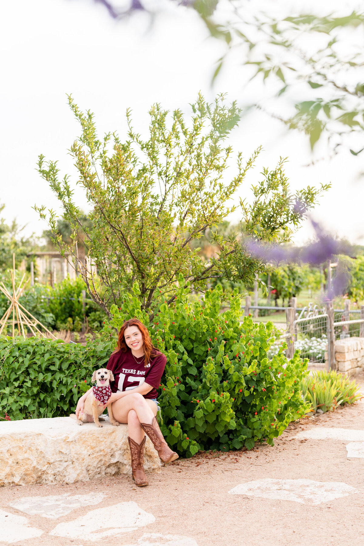 Texas A&M senior girl sitting on bench with her dog and wearing maroon jersey in middle of Leaching Teaching Gardens