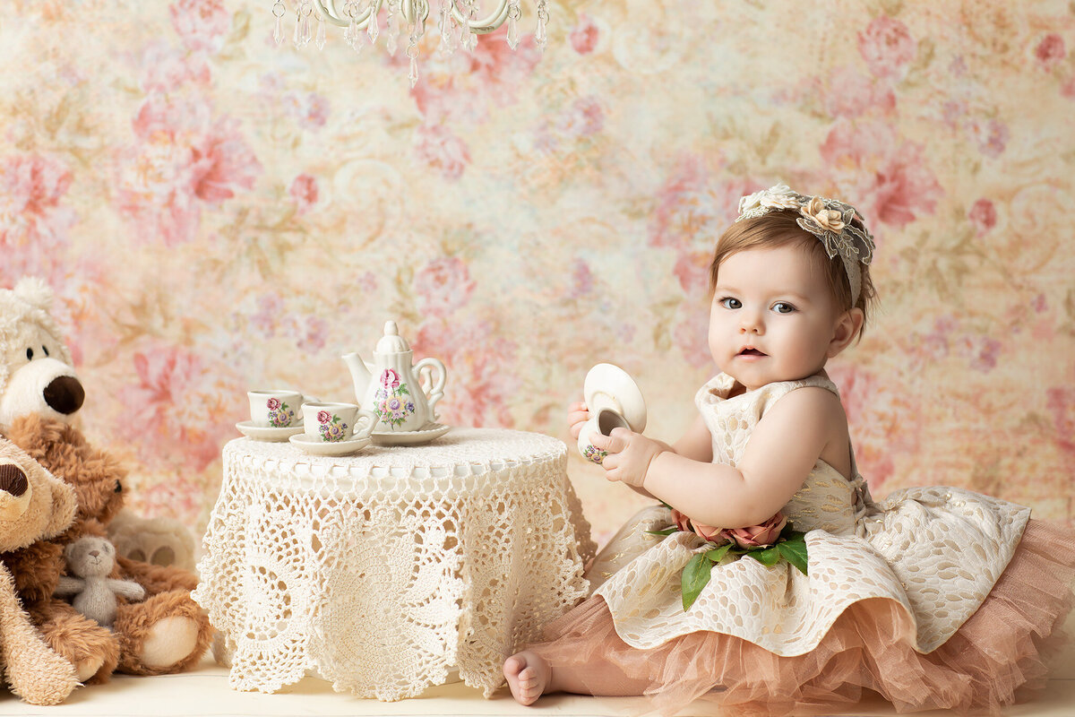 Baby girl in a gold and blush dress with a floral headband holds a tiny teacup at a lace-covered table set for tea, surrounded by plush teddy bears and delicate china, photographed by a Dallas milestone photographer.