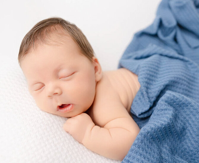 Baby newborn boy sleeping with blue waffle blanket over him while lying on white posing blanket.