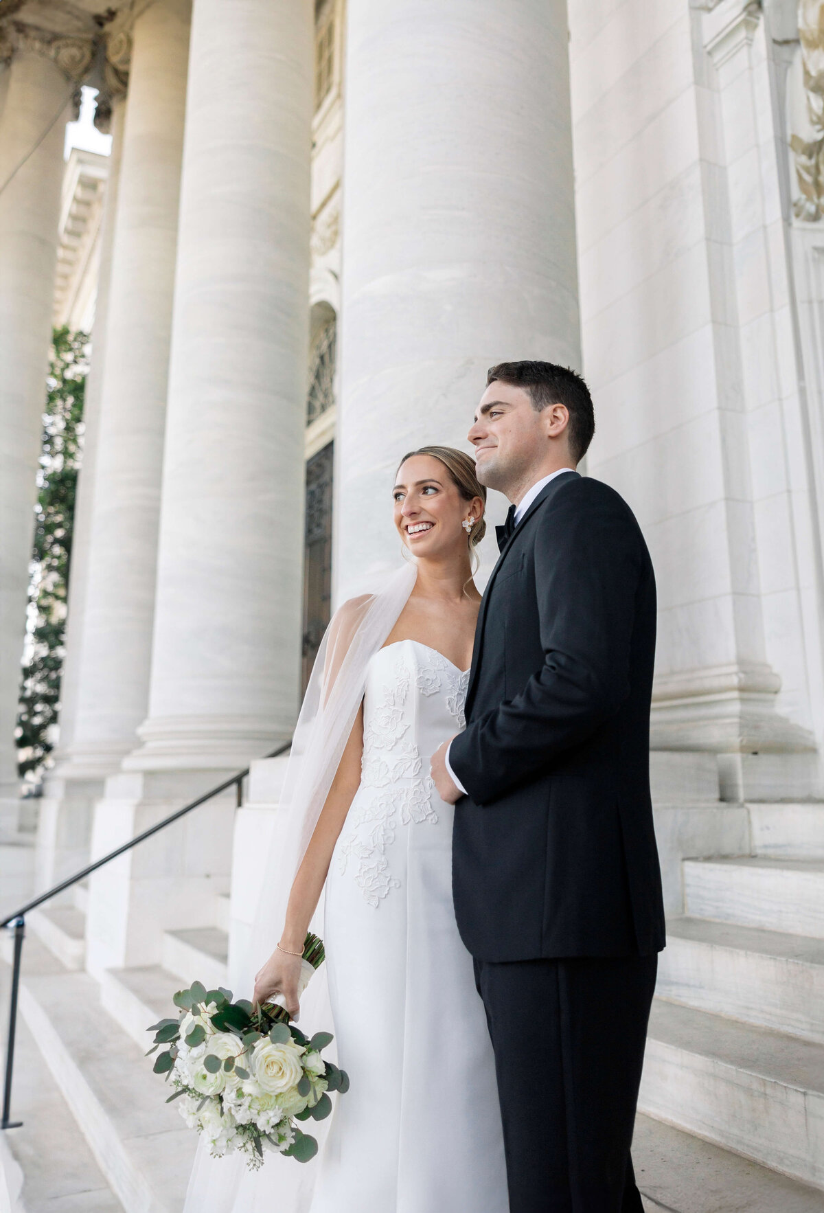 A bride and groom stand side by side on marble steps, smiling. The bride holds a bouquet of white roses and greenery, wearing a white dress and veil. The groom is in a black suit. Tall columns appear in the background.