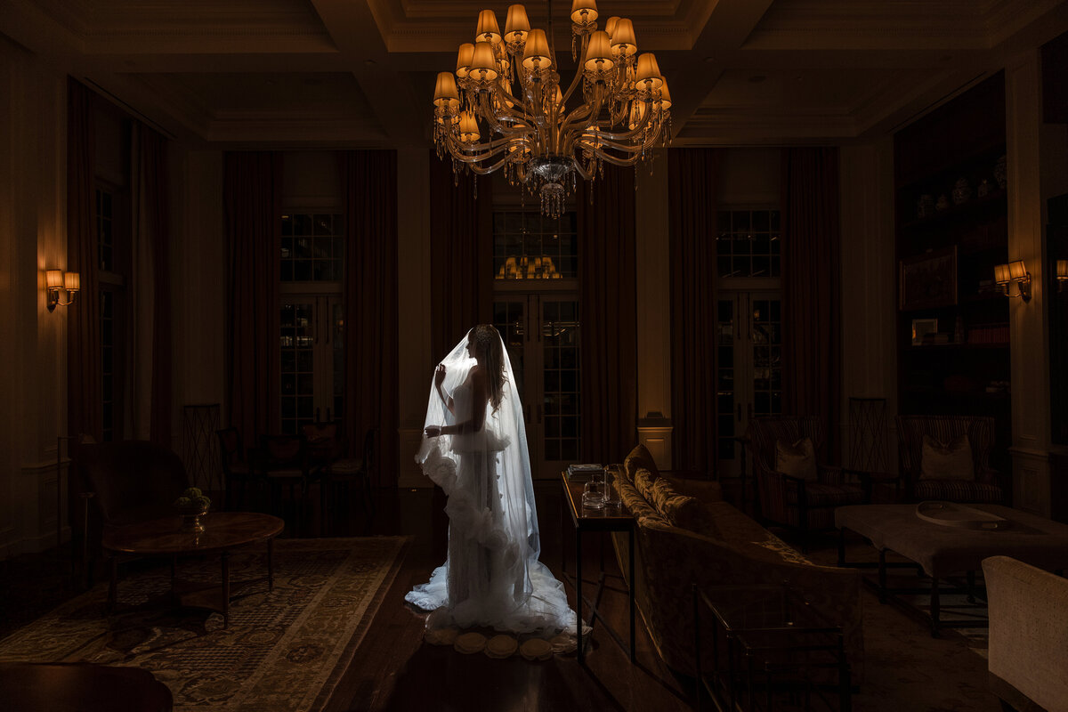 A silhouetted wedding portrait of a person in a long veil and gown, standing in the center of a dark, elegant room under a glowing chandelier, with warm light from sconces and reflections softly outlining the surrounding furniture.