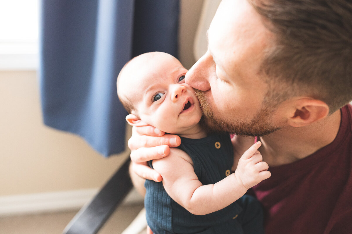 Dad kissing newborn baby's cheek in nursery. Image by Portland Oregon newborn photographer.