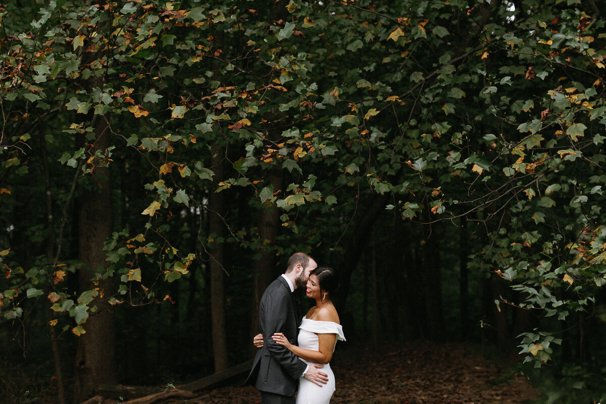 Bride and groom sharing intimate embrace under a tree with green leaves starting to turn orange. The lighting is natural and moody.