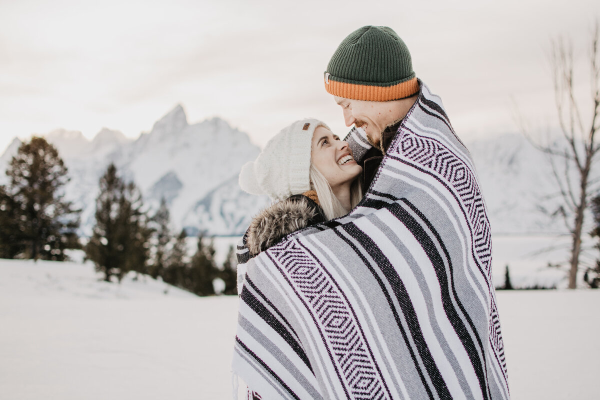 man holding woman while standing in a snowy field in Jackson Hole, as they are wrapped up in a striped blanket photographed by jackson hole photographers