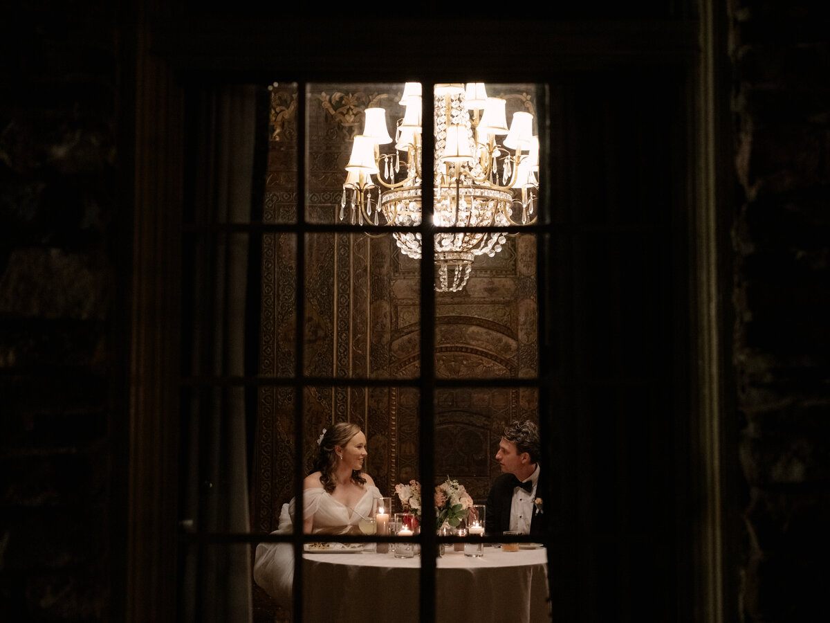 November wedding at Graylyn Estate, designed and planned by Charming Carolina — intimate moment captured through the window as bride and groom share a private dinner and laugh together in the Persian Card Room of the manor house.