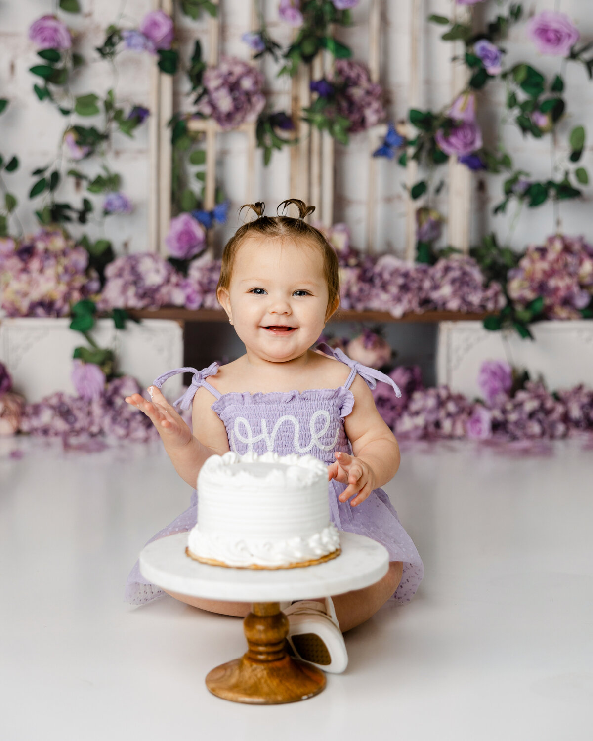 Smiling baby in a lavender “one” dress during a cake smash with purple flowers in the background. Photograph by Yucaipa cake smash photographer Kaitlyn Dawn Photography.