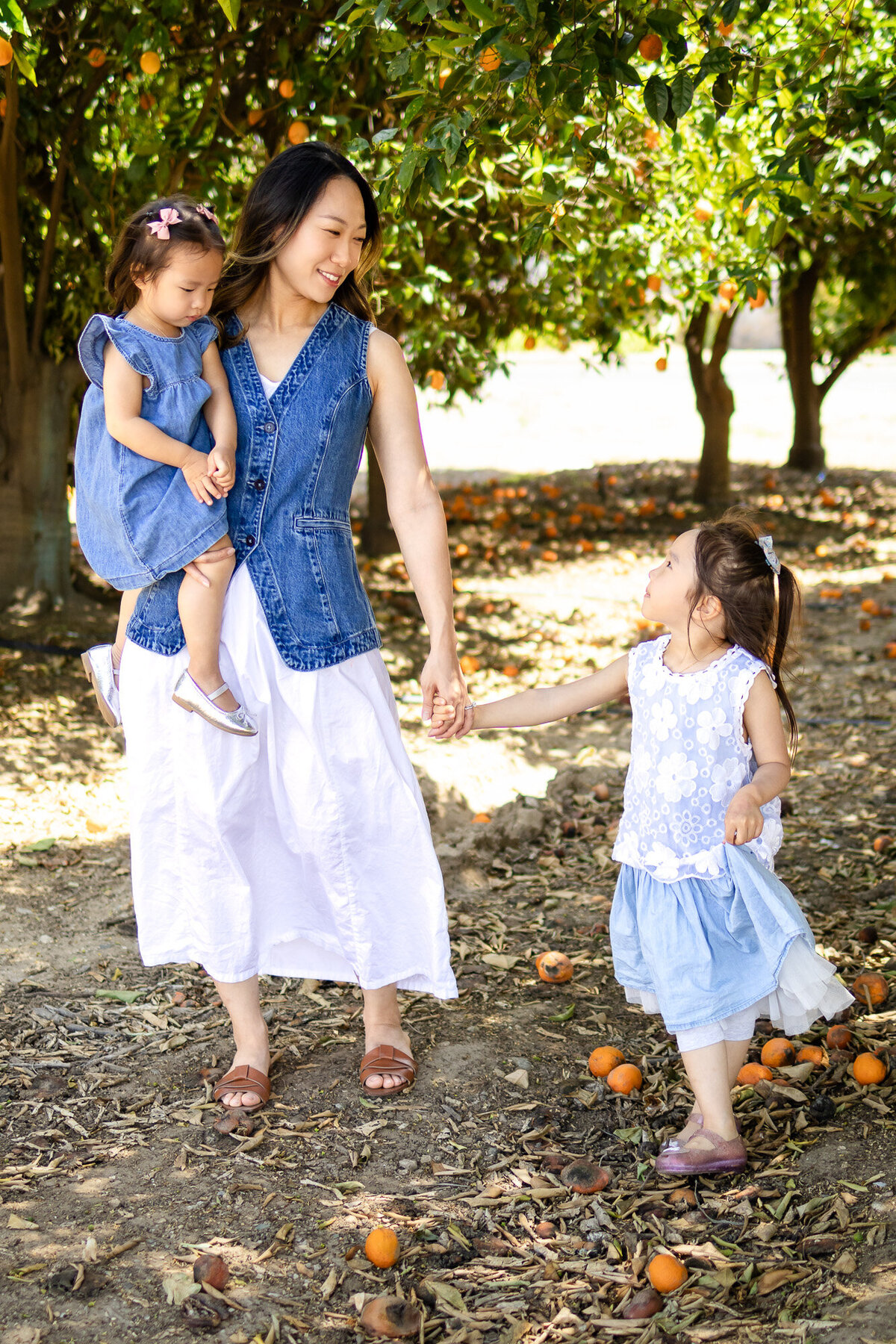 Bay Area Family Portfolio - Ellobelle Photography captures a mother holding hands with one daughter and carrying another in a sunlit orchard surrounded by fallen oranges.