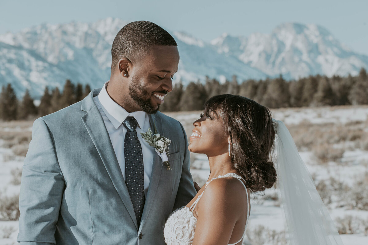 bride looking up to her groom as he smiles down at her with the snowy Tetons in the distance for their winter elopement