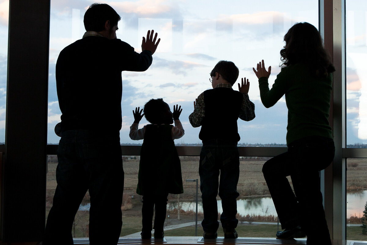 a family of four presses hands against window in a silhouette photo