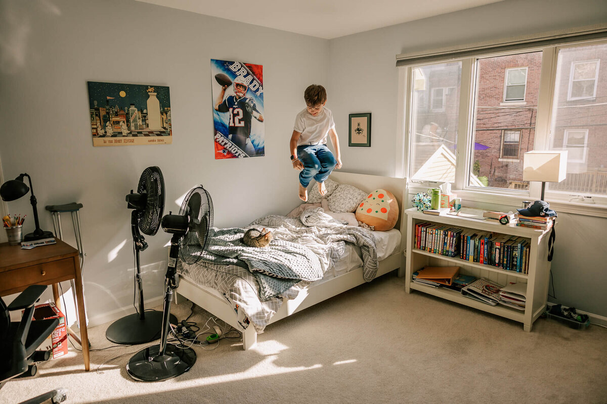 Young child is captured mid-air with his knees bent, hovering over his bed in the sunshine. A small grey cat is curled up at the foot of the bed, looking up at him curiously.