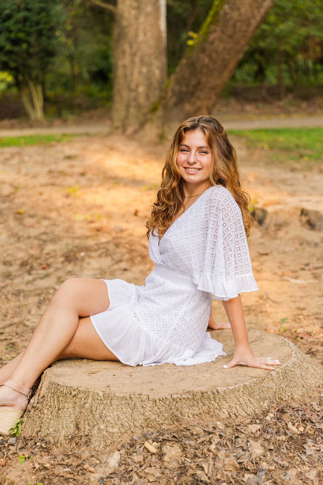 senior wearing a white texted short dress sitting on a tree trunk legs crossed during portrait session