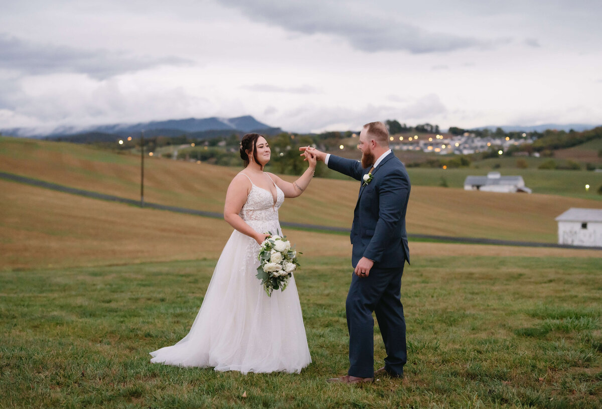 bride and groom dancing in a field for their Shenandoah wedding with man twirling his bride in circles on top of a hill with a town in the distance