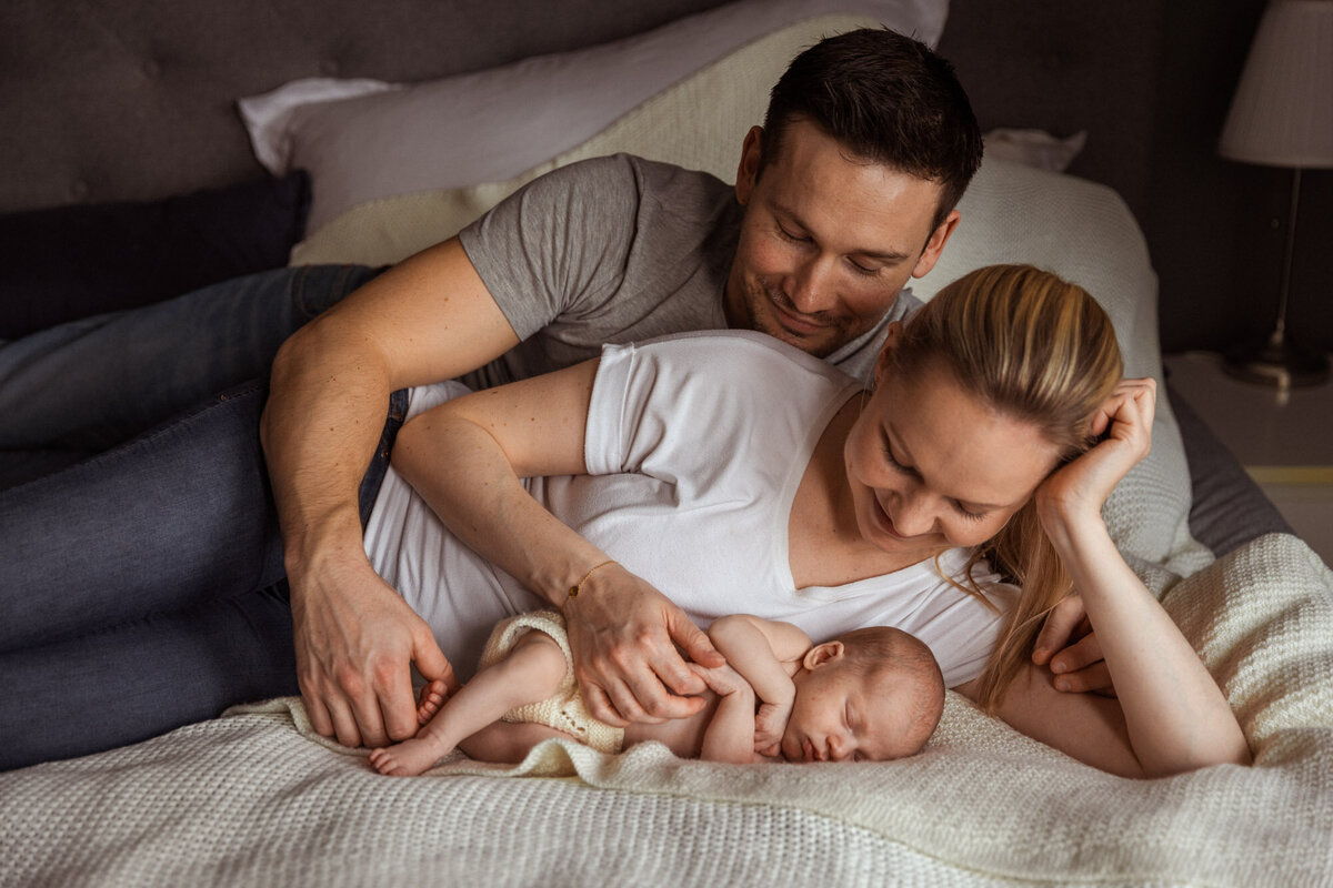 Livsstilsfoto familiefoto av foreldre som ligger på en seng og ser på babyen sin. Far er bakerst og holder rundt mor som holder hånden sin på babyen.