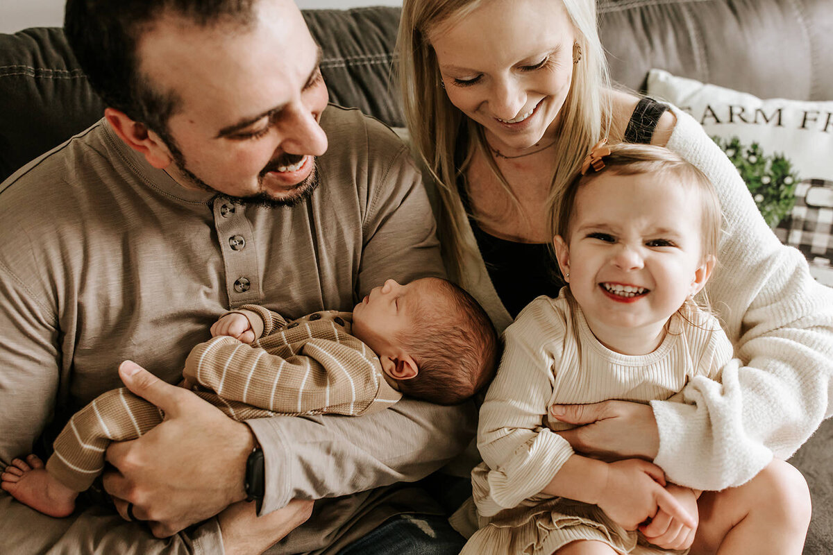 mom and dad smiling at toddler who is cheesing at the camera in a cozy white dress, while dad holds newborn baby cuddled in his arms.